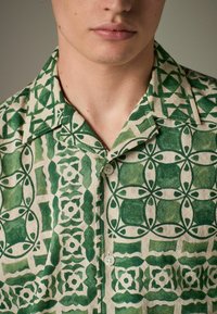 Chemise à manches courtes en tissu naturel, avec un motif géométrique vert sur un fond clair, dotée d'un col et de deux boutons.