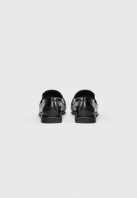 Mocasines de cuero negro con punta redondeada y tacón bajo, que presentan una textura suave y detalles de costura sutiles en la parte trasera.