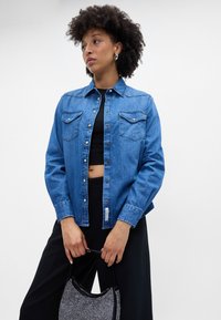 Camisa de ganga azul clara com botões e dois bolsos no peito, fechos metálicos e mangas compridas. O modelo combina-a com calças pretas e uma mala texturizada.