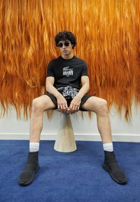 Černé bavlněné tričko s bílým logem Lacoste, vzorované šortky, tmavé vysoké tenisky a bílé ponožky, sedící na dřevěném stoličku. Oranžové pozadí.