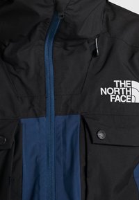 Veste noire et bleu marine avec fermeture éclair à l'avant, poches à boutons et logo blanc "The North Face" sur la poitrine.
