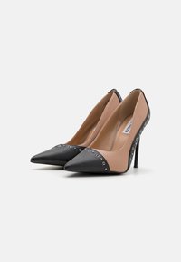 Beige og svarte høyhælte pumps med spiss tå, med nagledekorasjoner langs sømmen og en smidig, glatt finish.