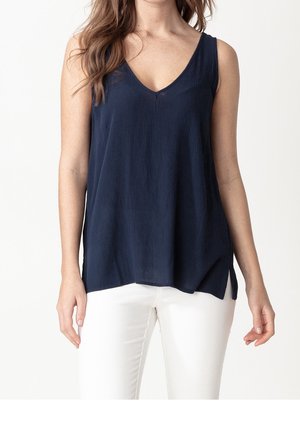 Nainen, jolla on yllään hihaton, laaja tummansininen V-kaula-aukollinen paita ja valkoiset housut, seisoo yksinkertaisen valkoisen taustan edessä.