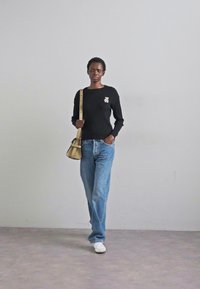 A model walks confidently, wearing a black sweater with a patch, light blue jeans, and white sneakers, carrying a small beige handbag.