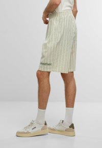Shorts rayés verts et crème avec une taille élastique, dotés de poches latérales et d'un logo sur la jambe. Portés avec des baskets blanches et des chaussettes.
