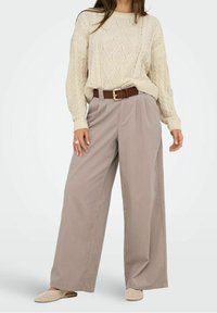 Hellbeiger Pullover mit Zopfmuster und Rundhalsausschnitt. Locker sitzende, hoch taillierte, hellgraue Hose mit weitem Bein, gesichert durch einen braunen Gürtel. Beigefarbene Slipper.