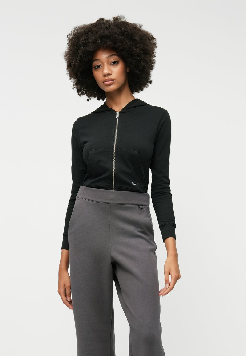 Hoodie noir zippé avec une poche avant, associé à un pantalon de jogging large gris avec des poches latérales et une boucle à la taille.