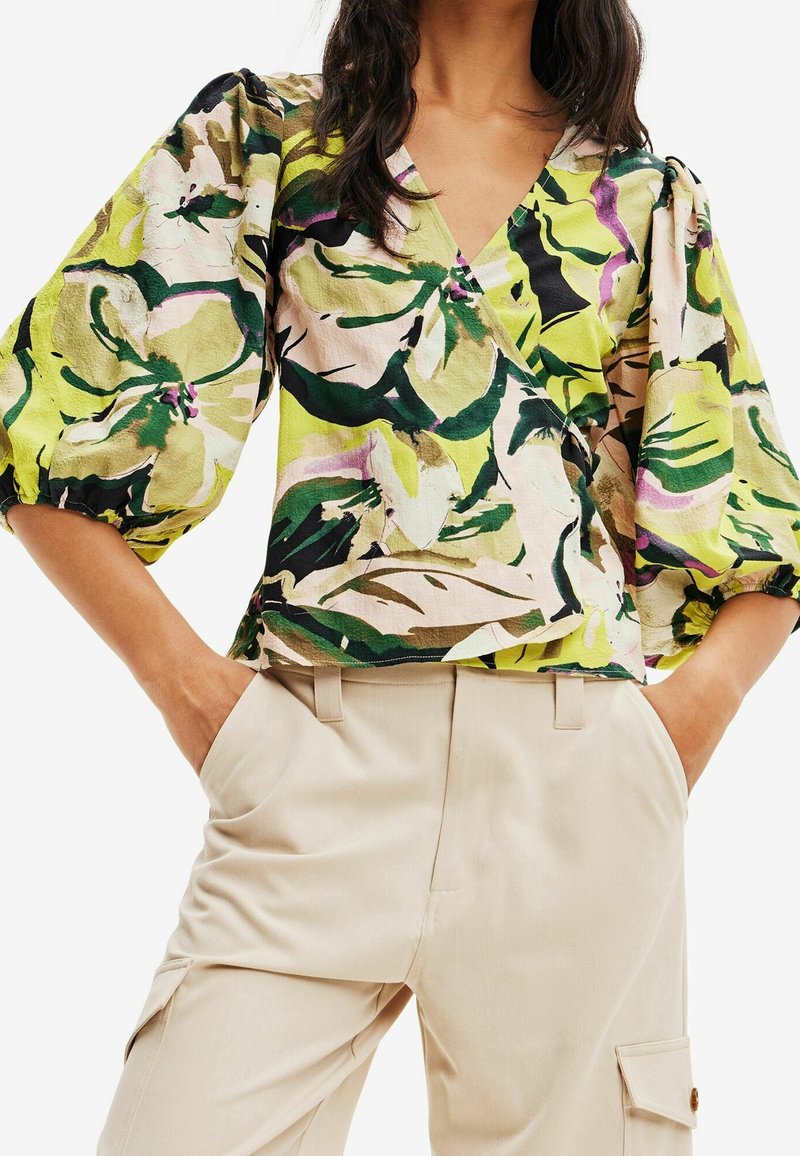 Femme portant un chemisier cache-cœur vert à motif floral avec des manches bouffantes et un pantalon beige taille haute, les mains dans les poches.