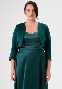 Abito di raso verde smeraldo con corpino aderente, scollatura quadrata e gonna fluida. Accompagnato da un giubbotto corto abbinato con maniche a tre quarti.