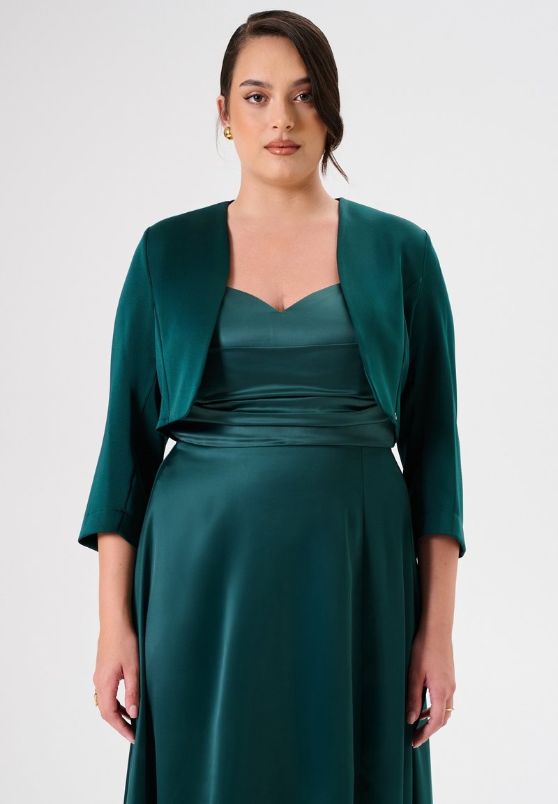Abito di raso verde smeraldo con corpino aderente, scollatura quadrata e gonna fluida. Accompagnato da un giubbotto corto abbinato con maniche a tre quarti.