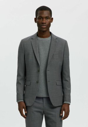 Homme portant une veste de costume gris ajustée et un pantalon assorti sur un pull gris côtelé, debout devant un fond clair uni.