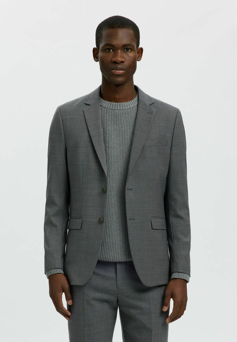 Homme portant une veste de costume gris ajustée et un pantalon assorti sur un pull gris côtelé, debout devant un fond clair uni.