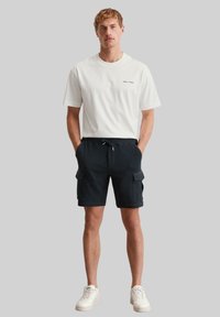 T-shirt en coton blanc avec un petit logo, associé à un short en cargo bleu marine comportant deux poches latérales et une taille à cordon. Baskets blanches.