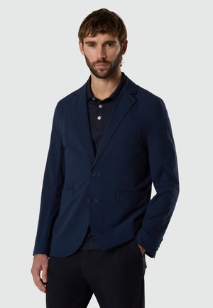 Uomo con barba che indossa un blazer blu scuro sopra una polo blu navy, in piedi davanti a uno sfondo grigio chiaro uniforme.