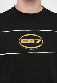 Juodas medvilninis marškinėliai su apvalia iškirpte, baltais horizontaliais dryžiais ir auksiniu ovaliu logotipu, kuriame užrašyta "EA7" ir "Emporio Armani".