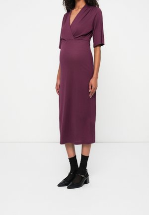 Femme portant une robe portefeuille prune longueur genou avec manches aux coudes, chaussettes noires et chaussures Mary Jane noires à talons bas sur un fond blanc uni.