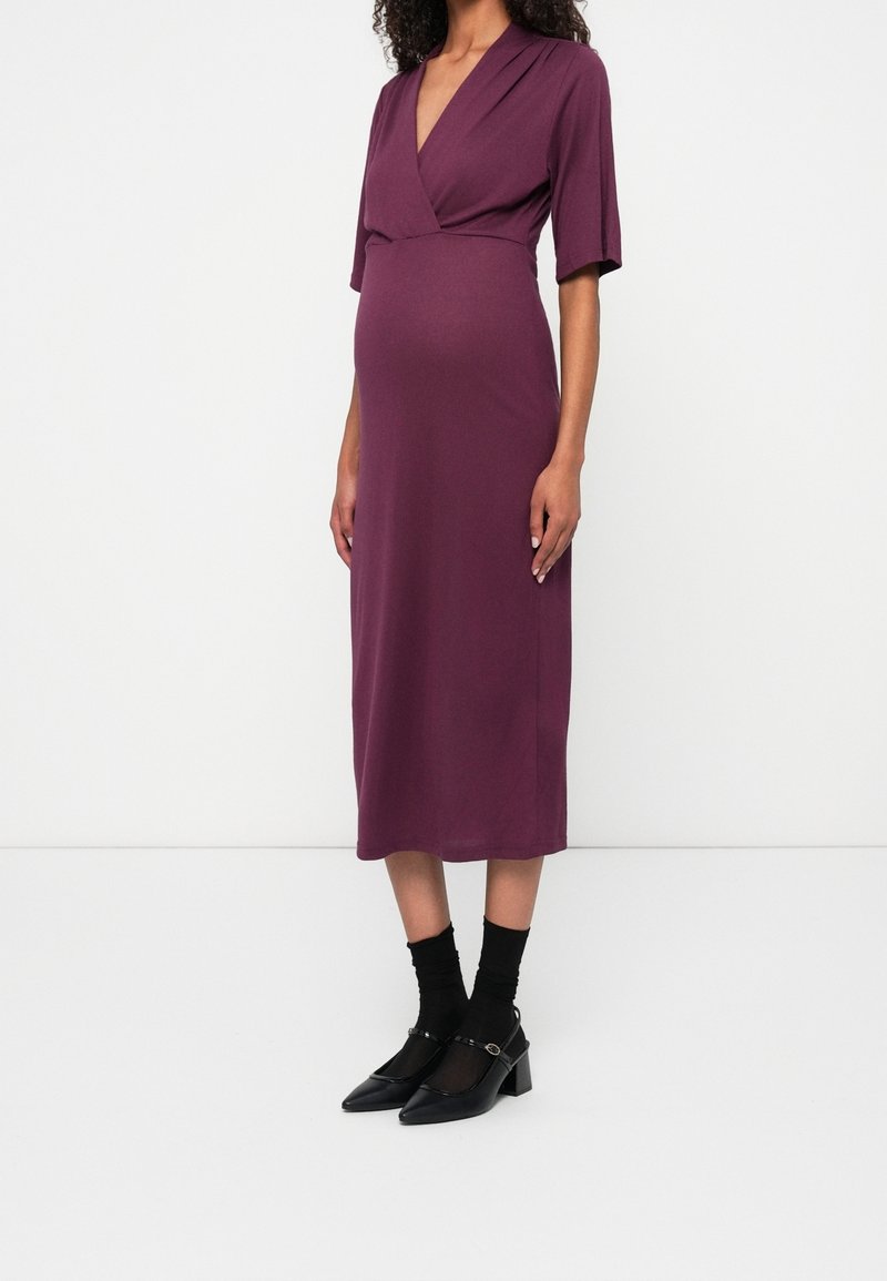 Femme portant une robe portefeuille prune longueur genou avec manches aux coudes, chaussettes noires et chaussures Mary Jane noires à talons bas sur un fond blanc uni.