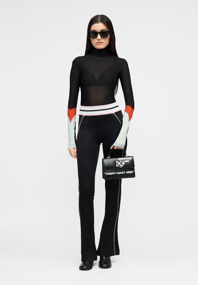 Γυναίκα που φοράει μαύρη ημιδιάφανη μπλούζα με μακριά μανίκια, μαύρο παντελόνι με λευκή ρίγα, μαύρες μπότες, γυαλιά ηλίου, κρατώντας μία μαύρη τσάντα με λευκό κείμενο.