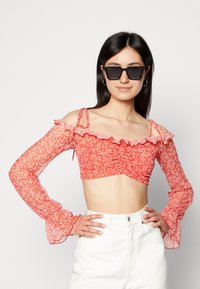 Top curto floral vermelho com mangas longas transparentes, decote com babados e detalhe de laço nos ombros, combinado com calças brancas de cintura alta.