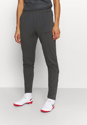 Personne portant un pantalon de sport noir Nike et des crampons blancs Nike avec des crampons rouges, debout devant un fond clair uni.