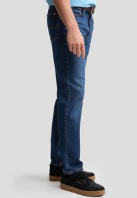Blaue Denim-Jeans mit geradem Bein, kombiniert mit einem hellblauen Hemd und schwarzen Sneakers mit Gummisohle. Schlichte Nahtakzente.