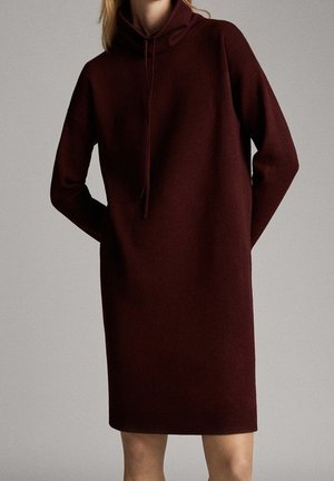 Femme portant une robe tricotée marron foncé à manches longues, longueur genou, avec une capuche et un cordon de serrage, debout devant un fond uni.