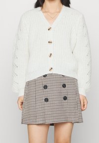 Vit stickad cardigan med v-ringad hals, utsmyckad med stora knappar och texturerade ärmar, tillsammans med en brun rutmönstrad kjol med svarta knappar.