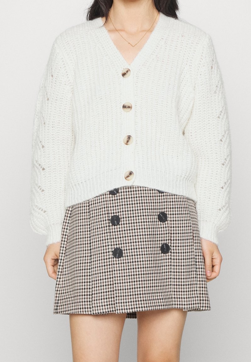 Vit stickad cardigan med v-ringad hals, utsmyckad med stora knappar och texturerade ärmar, tillsammans med en brun rutmönstrad kjol med svarta knappar.