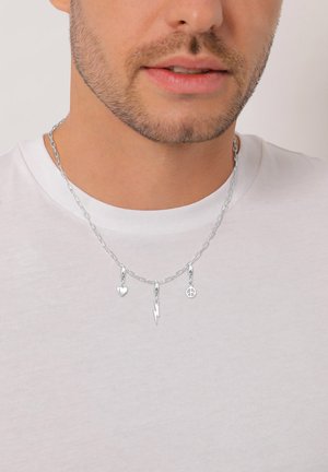 Ein Mann trägt eine silberne Halskette mit Anhängern in Form eines Herzens, eines Blitzes und eines Friedenssymbols über einem weißen T-Shirt.
