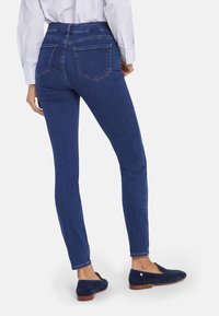 Jeans skinny azul oscuro con una textura suave, de talle alto y detalles de costura sutiles. Combinados con mocasines navy sobre un fondo blanco.