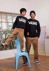 Twee personen dragen zwarte Vans-hoodies met een wit logo, khaki broeken en zwarte Vans-schoenen. Er wordt een blauwe metalen stoel gebruikt ter ondersteuning.