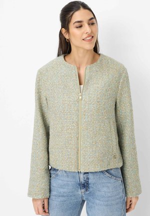 Frau trägt eine pastellfarbene Tweed-Jacke mit Reißverschluss und blaue Jeans, schaut leicht nach links und lächelt leicht.