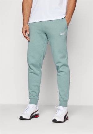 Hombre con pantalones deportivos Puma de color verde claro, zapatillas Puma blancas y una camiseta blanca, de pie con una mano en el bolsillo.