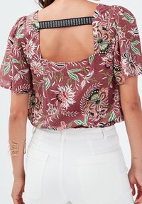 Rote Blumenbluse mit kurzen Puffärmeln, offenem Rückendesign und gemusterten Details, kombiniert mit weißen hochsitztenden Hosen.