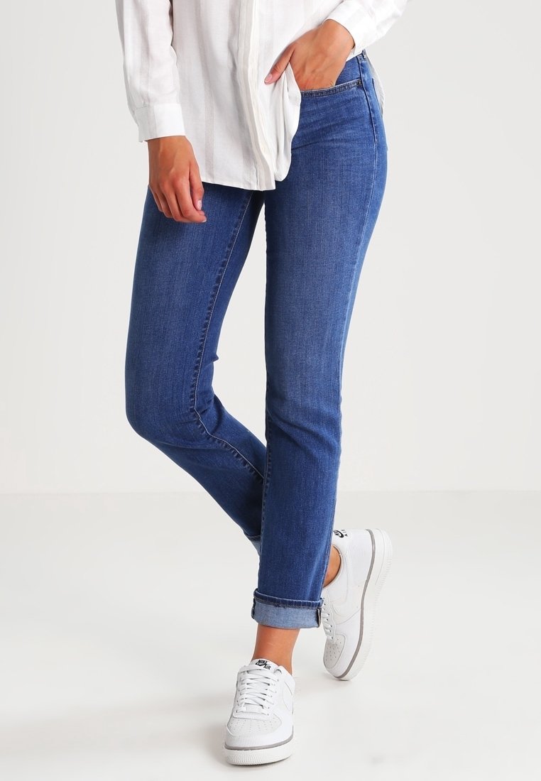 Partie inférieure d'une personne portant un jean bleu retroussé, des baskets blanches et une chemise blanche à manches longues, une main dans la poche, debout sur un fond uni.
