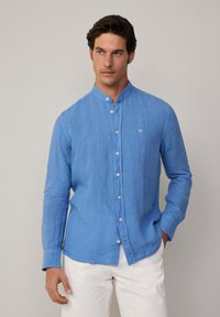 Camicia di lino azzurro chiaro con colletto a banda, maniche lunghe, chiusura a bottoni e un piccolo dettaglio ricamato sul petto.