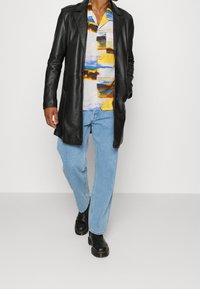 Svart läderkappa över en färgglad skjorta med abstrakt mönster; ljusblå jeans och svarta stövlar kompletterar outfiten.