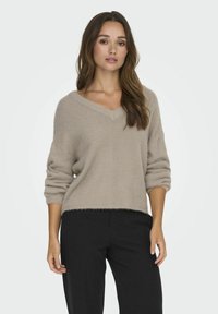 Beige, fuzzy trui met een v-hals en driekwart mouwen; heeft een losse fit en een franjerand, gecombineerd met zwarte broek.