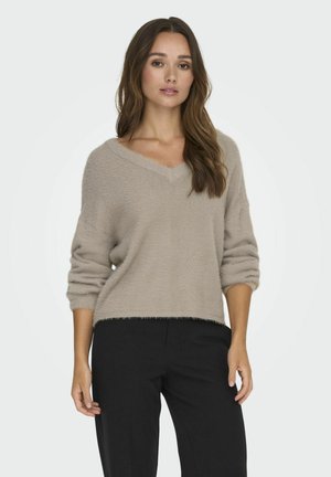 Beige, fuzzy trui met een v-hals en driekwart mouwen; heeft een losse fit en een franjerand, gecombineerd met zwarte broek.
