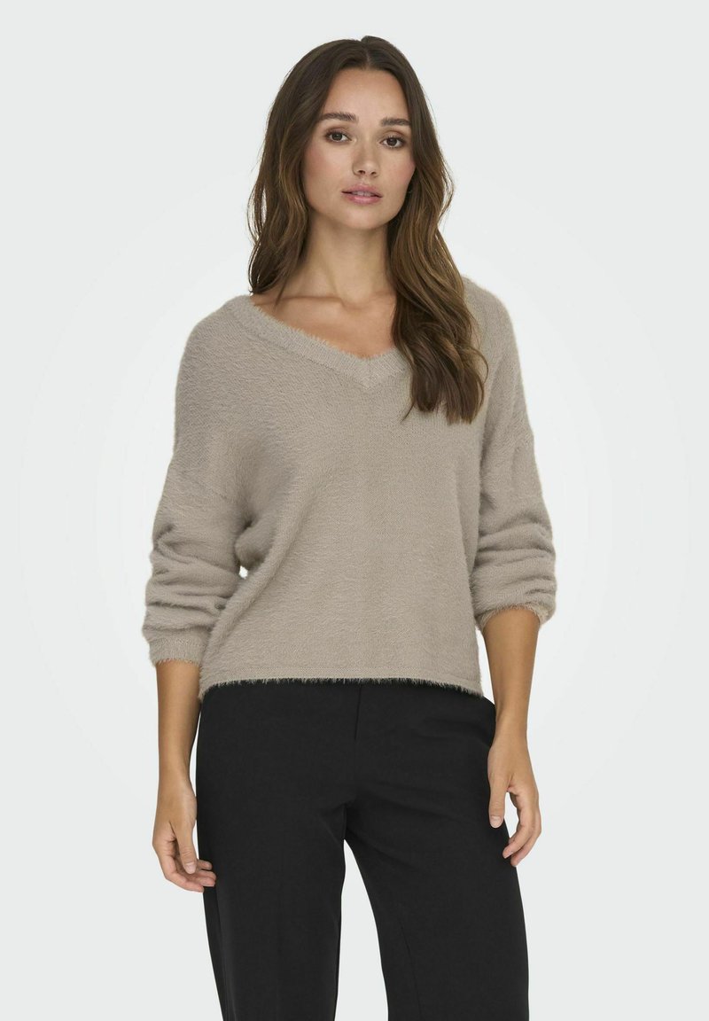 Pull beige moelleux à col en V et manches trois quarts ; présente une coupe ample et un ourlet effiloché, associé à un pantalon noir.