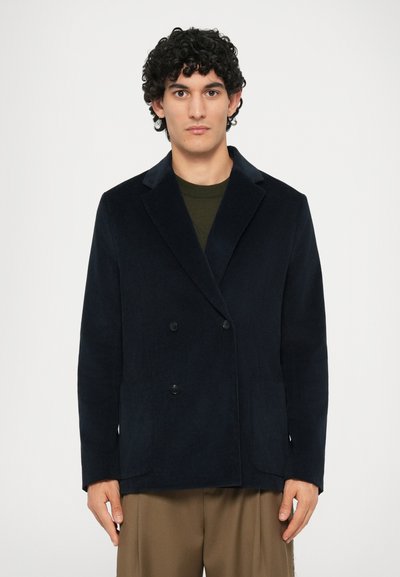 Veste croisée marine en tissu texturé, dotée de revers à crans et de deux poches avant. Portée sur un haut olive.