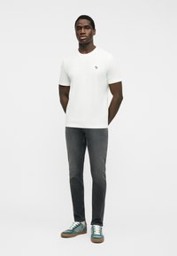 Hombre con camiseta blanca de manga corta lisa, jeans grises y zapatillas verdes, de pie con las manos detrás de la espalda sobre fondo blanco.