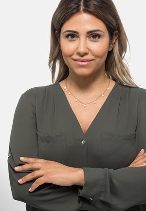 Goldfarbenes Necklace mit traumartigen Akzenten, getragen über einer dunkelgrünen Bluse mit zwei Brusttaschen und einer Knopfverschluss.