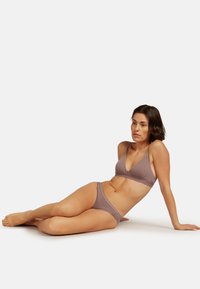 Donna sdraiata sul pavimento mentre indossa un reggiseno e mutandine color malva su uno sfondo chiaro e semplice.