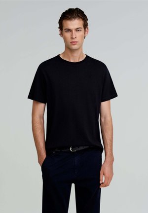 Camiseta de manga corta negra de algodón; cuello redondo, corte holgado, combinada con pantalones oscuros y un cinturón negro con hebilla plateada.