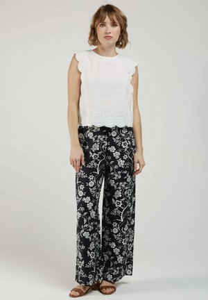 Femme debout portant un chemisier blanc sans manches à bord festonné et un pantalon noir à jambes larges avec des motifs floraux blancs, associé à des sandales marron.