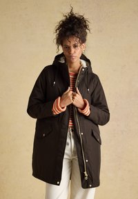 Parka noire à capuche avec une fermeture éclair, dotée d'une doublure douce, de deux poches avant et de poignets rayés orange, portée sur un pull rayé rouge et blanc.