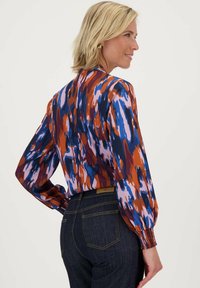 Multikleurige blouse met gedurfde patronen in blauw, oranje, roze en donkere tinten. Lange mouwen met gerimpelde manchetten en een gladde stofstructuur.