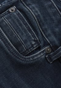 Tissu en denim bleu foncé avec une poche avant, détail de bouton en métal et coutures raffinées. La texture semble légèrement usée.
