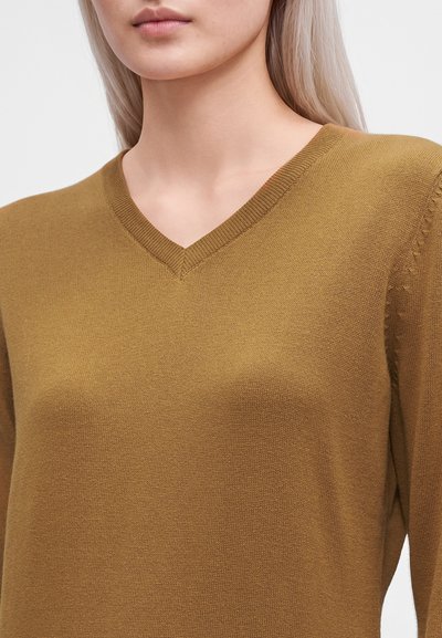 Femme portant un pull en maille jaune moutarde à col en V, montrant l'encolure et le haut du torse sur un fond uni.